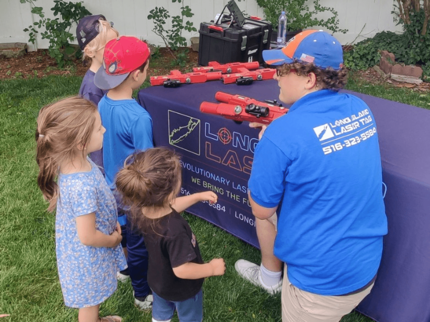 A Long Island Laser Tag employee leaning down to assist younger players with getting acquainted with their laser tag equipment. There are blue and red laser taggers on a table in the background of the image.